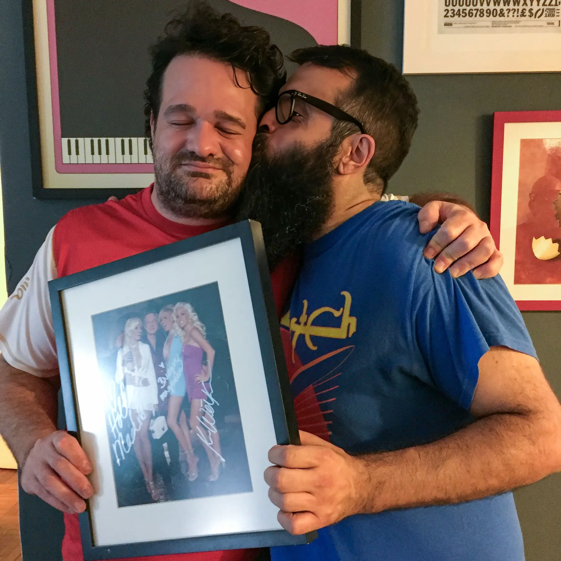 Jardel e Daniel Motta abraçados; Daniel beija o rosto de Jardel enquanto ele segura um quadro com uma foto emoldurada.