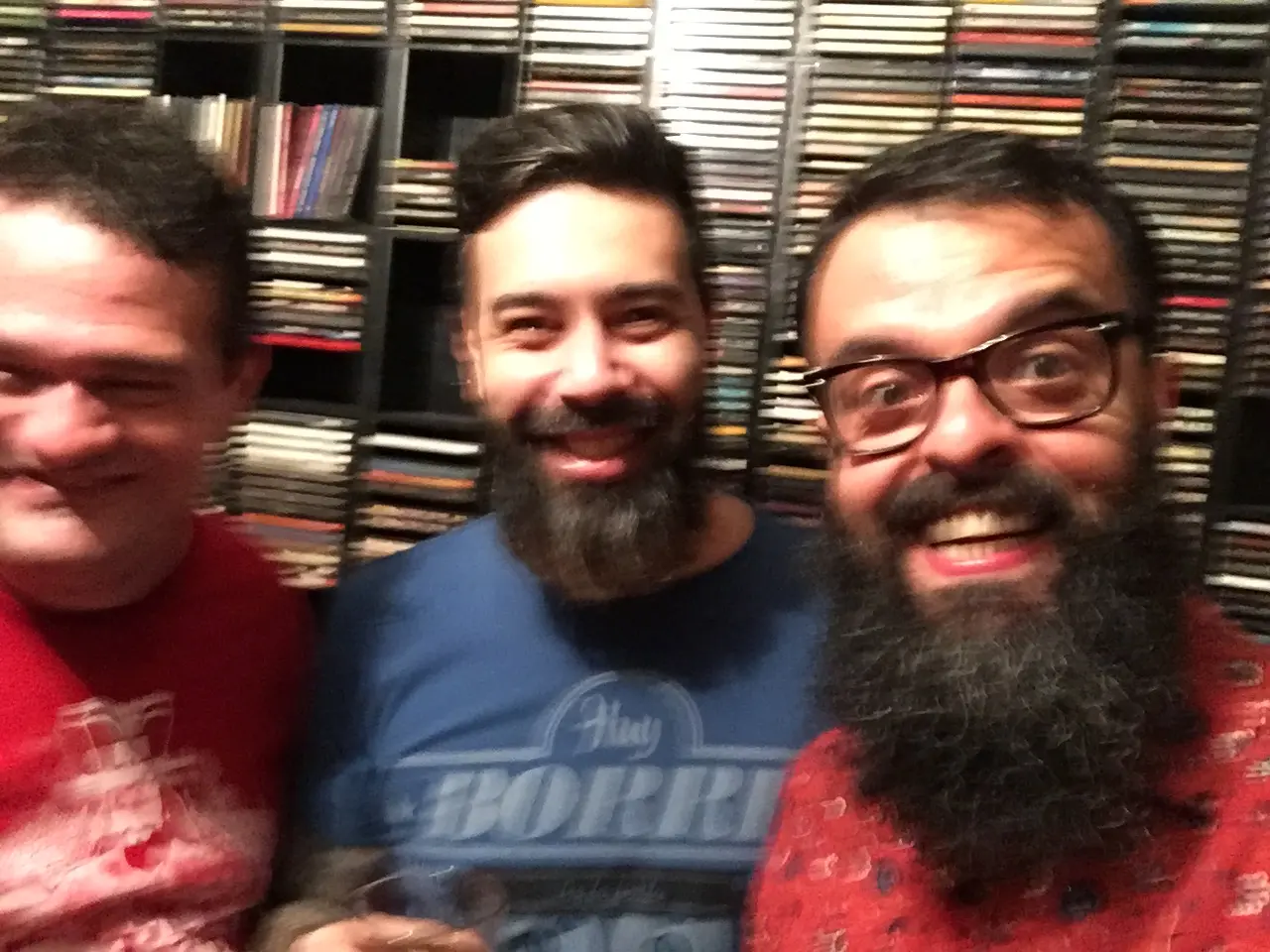 Jardel, Marco Bezzi e Daniel Motta sorrindo em um quarto com paredes completamente cobertas por porta-CDs.